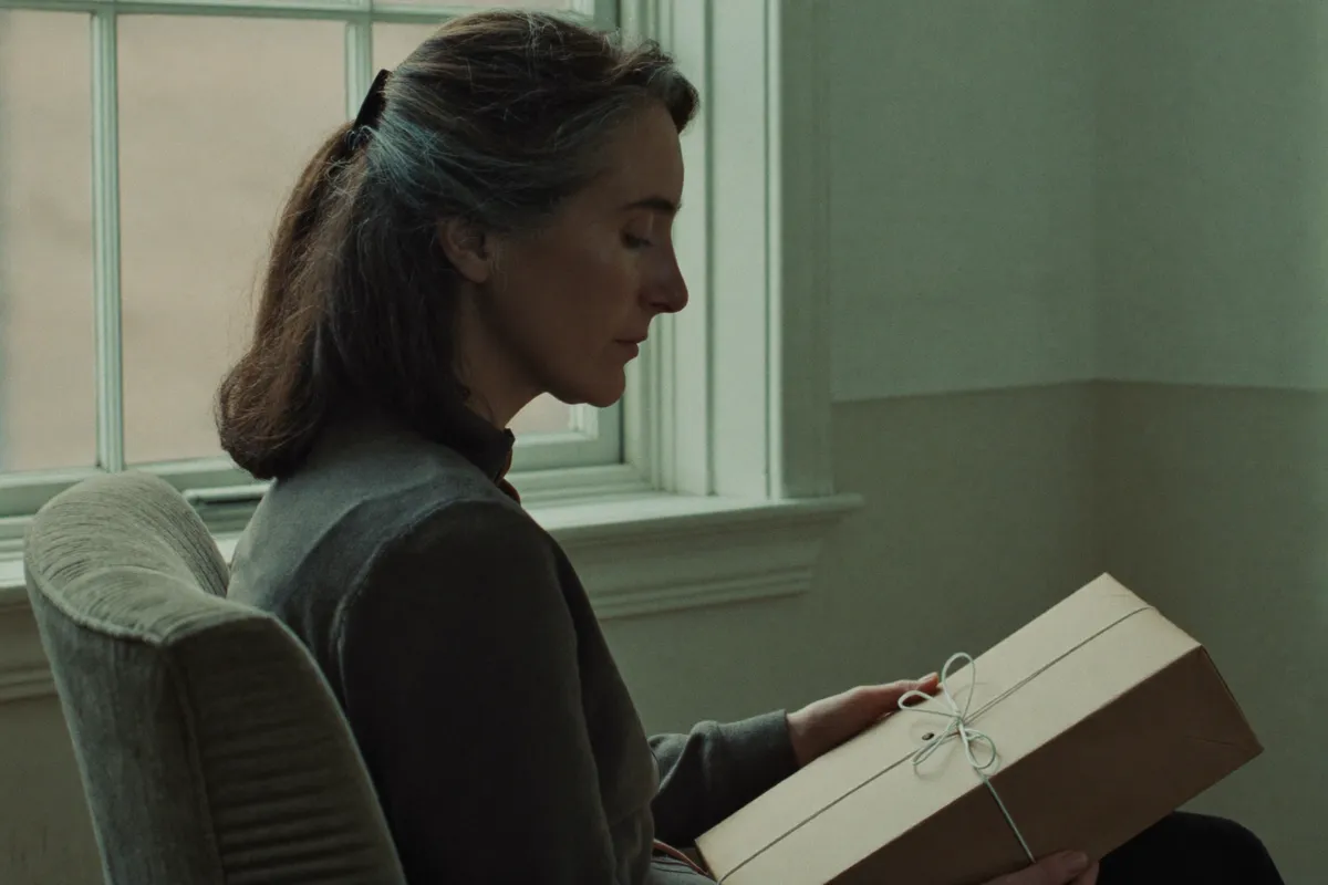 A woman in her forties by a window, looking down at a small gift in plain paper on her lap, the one talent she arrived with