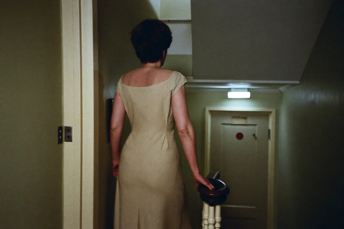 A woman at the foot of the stairs at night, looking up at a landing light still on, the default parent still on duty long after the children grew