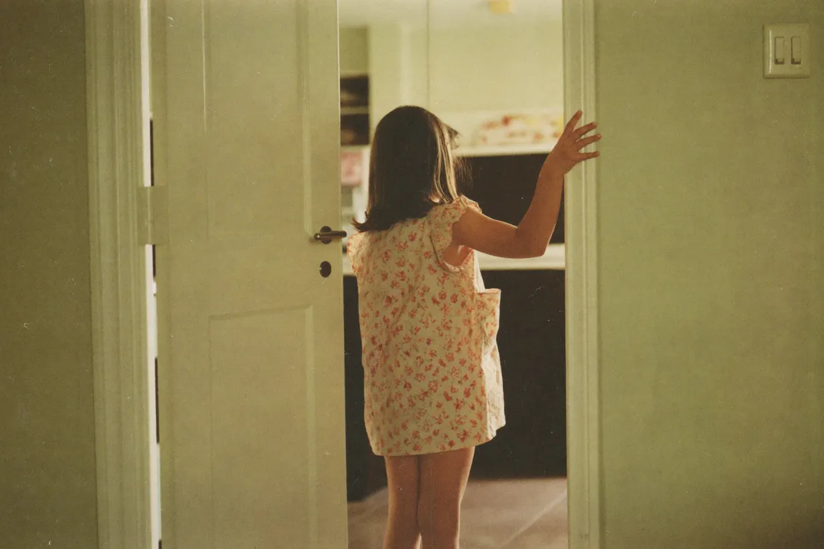 A young girl stands in a kitchen doorway with her back to us, reading the room before she walks into it, the earliest sign of eldest daughter syndrome and parentification