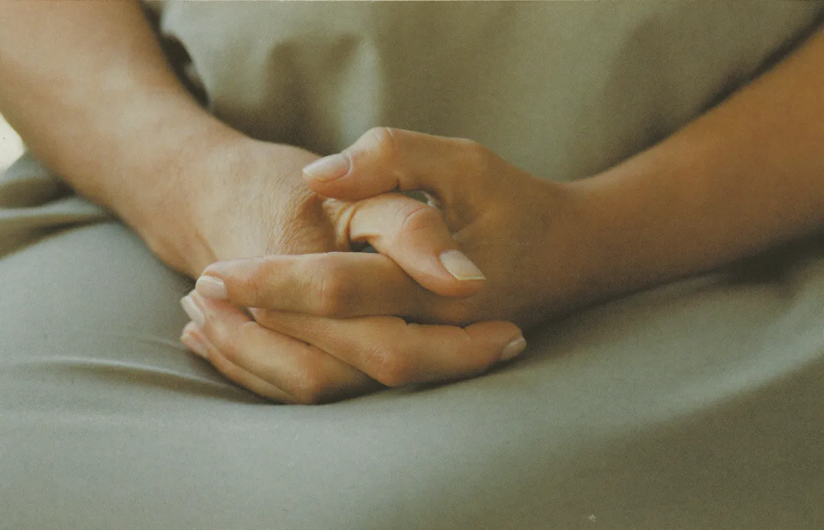 A woman's hands clasped tightly in her lap, holding a need back rather than asking for anything