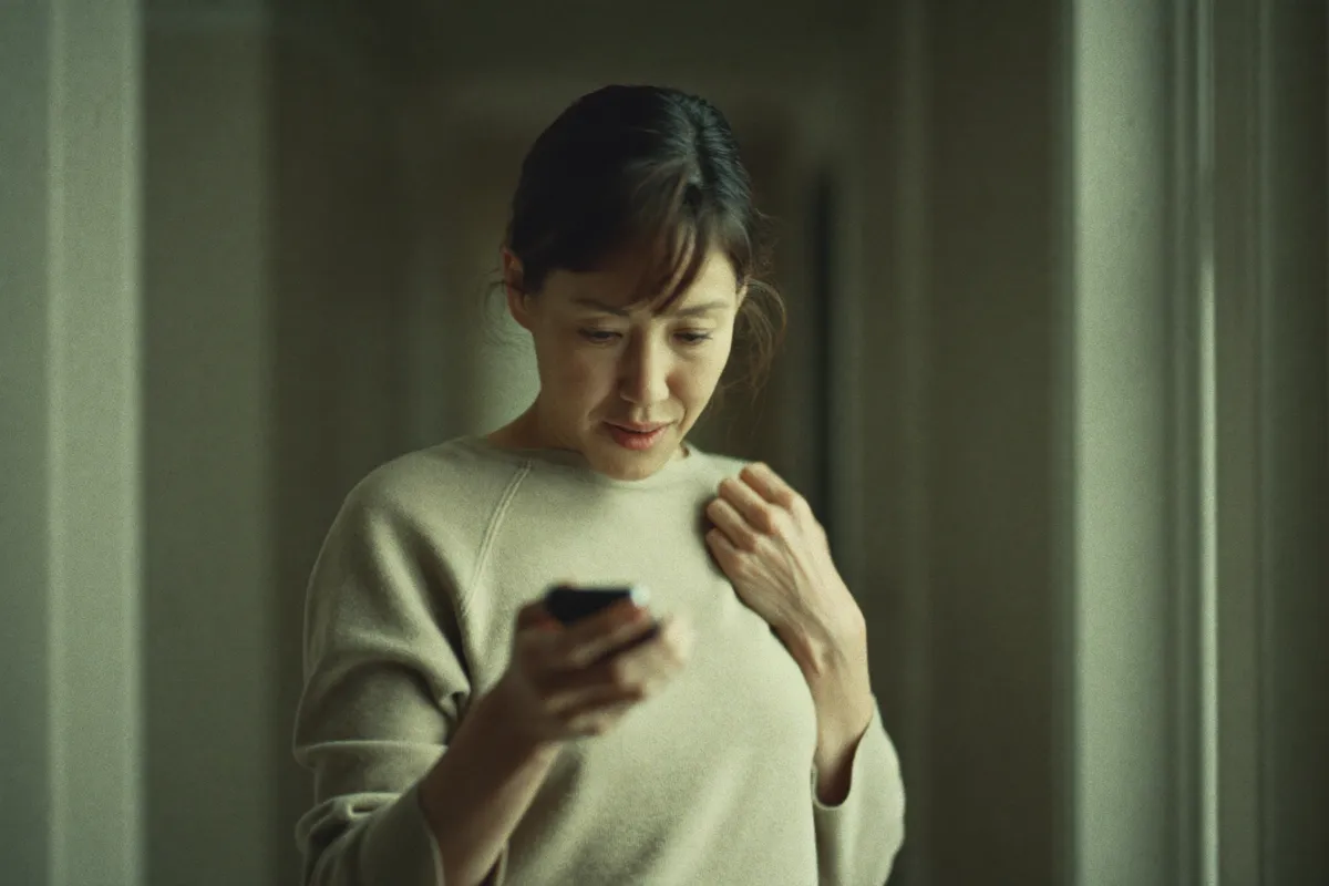 A woman in her forties alone in a hallway, looking down at her phone with one hand at her collarbone