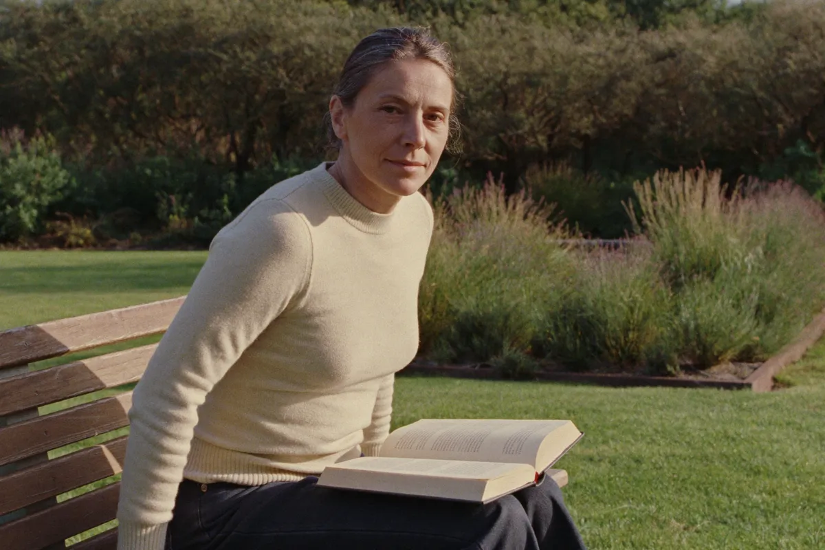 A woman in her forties on a garden bench with an open book in her lap, looking up from the page