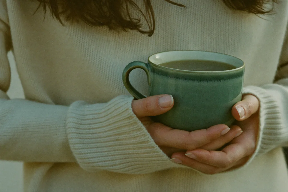 A woman in a knit sweater holding a warm mug of tea in both hands, a small grounded sensory moment
