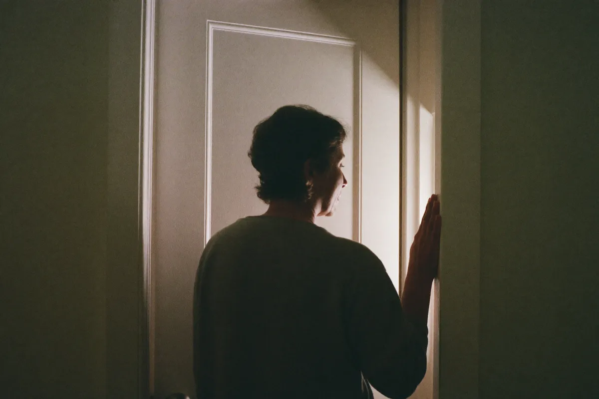 A woman standing still in her own hallway, hand against the wall, head turned toward a sound she has already read, the everyday shape of hypervigilance