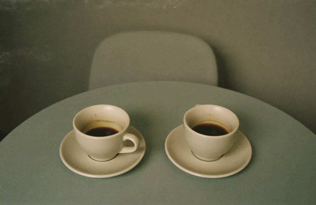 A woman sitting alone at her kitchen table at night, phone face down beside a cold cup of tea · the quiet reality of making friends in midlife