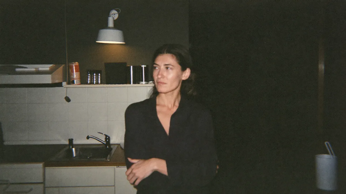 A woman standing in her own kitchen at dusk like a guest in her own home, the midlife feeling of not recognizing your life