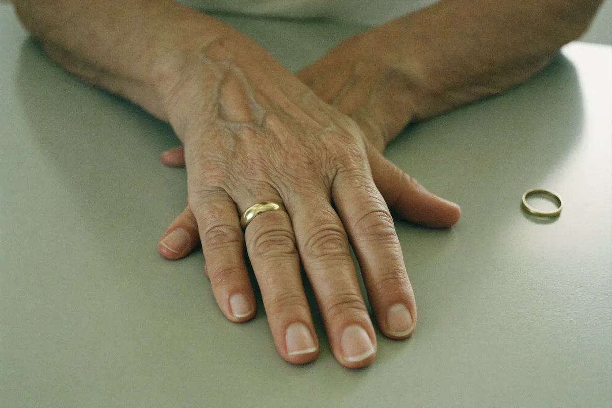 A woman's hands on a kitchen table, one wedding ring still worn and an older ring set down beside it, the married name and the birth name