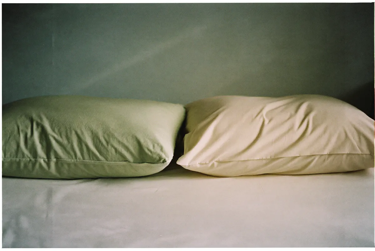 A woman sitting alone on the edge of the bed at dawn, her partner asleep behind her, showing the quiet distance of perimenopause and relationships