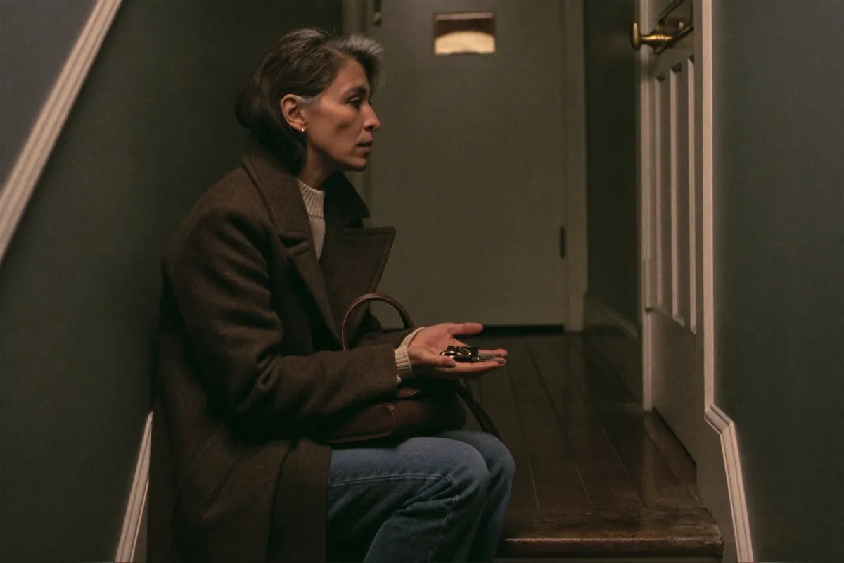 A woman in her forties sitting on the bottom stair in her coat, keys in her hand, having decided not to go out after all