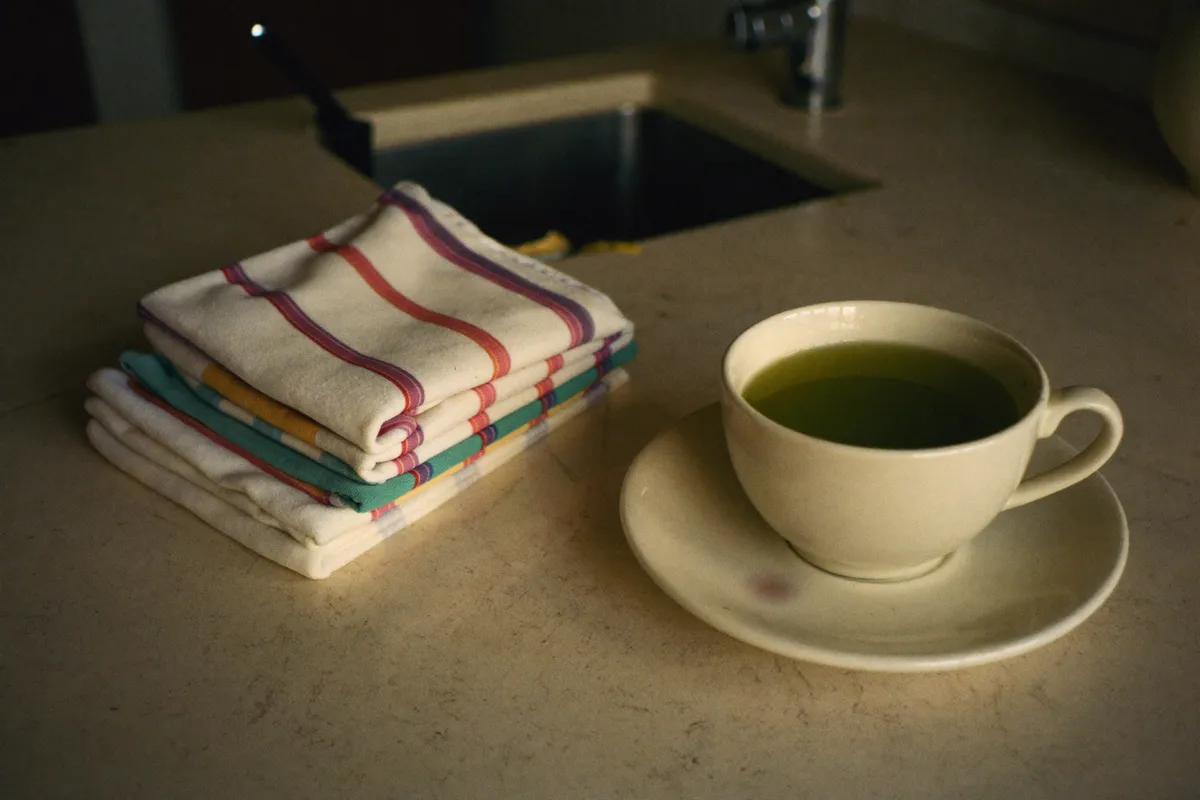 A woman sitting alone at a kitchen table in the early morning, hands around a cup she has not drunk from, the resentment of being needed sitting quietly with her