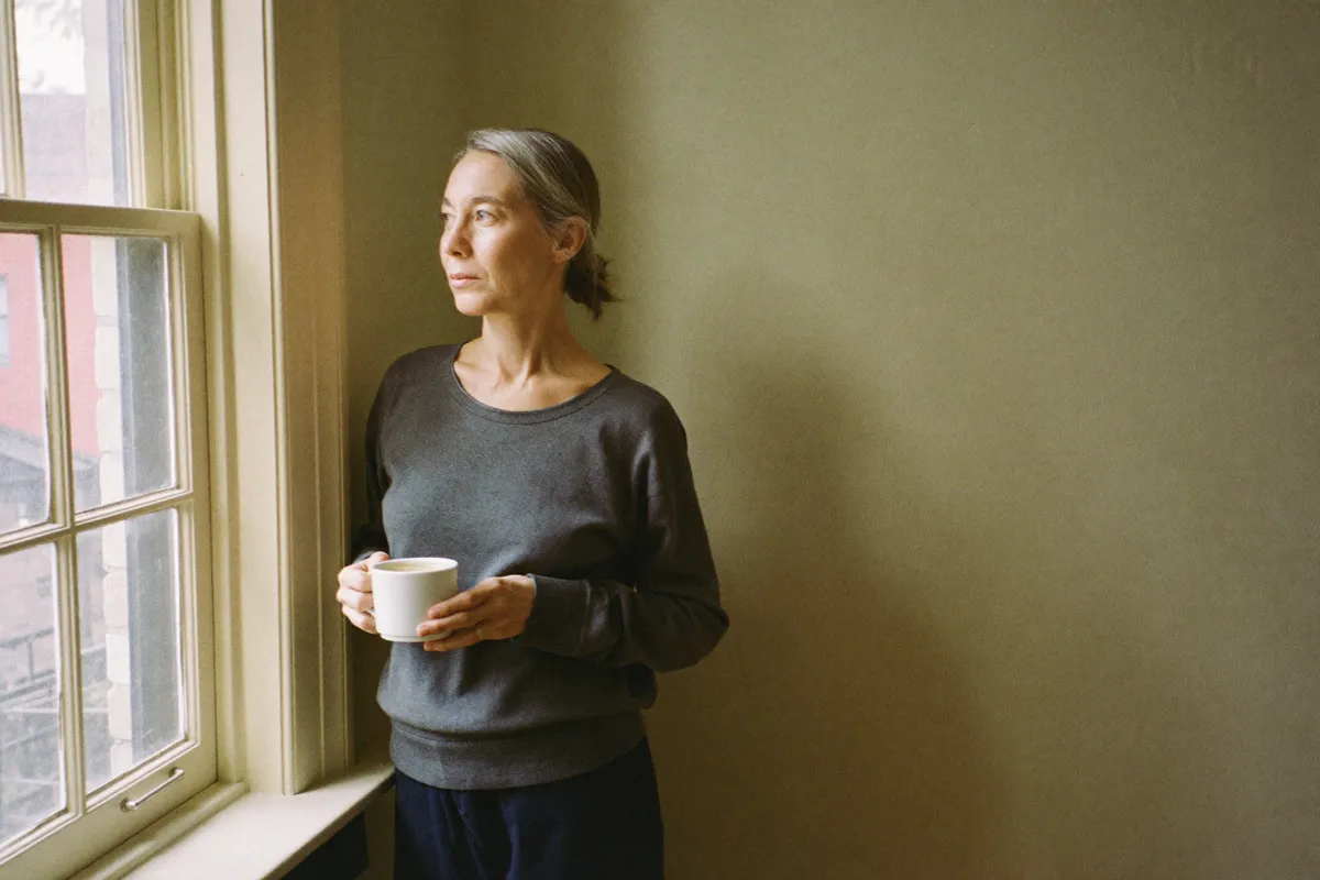 A woman stands at her kitchen window in the early morning with a mug in both hands, taking the small protected time that makes sandwich generation caregiving survivable