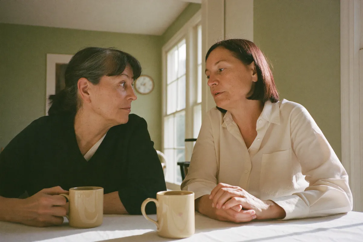 Two women sit close at a kitchen table in the afternoon, one listening properly while the other speaks, because strong friends need friends too