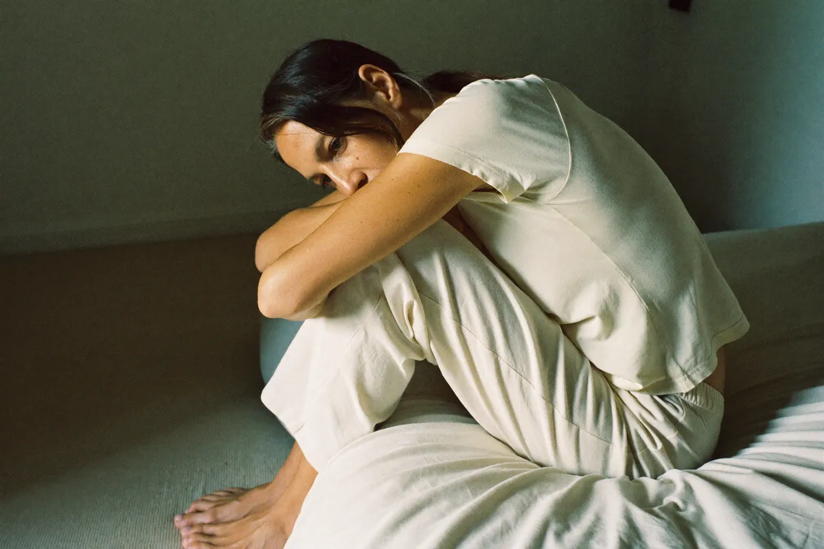A woman in her forties sitting on the bed in the early light with her knees drawn up, gathering herself before the day